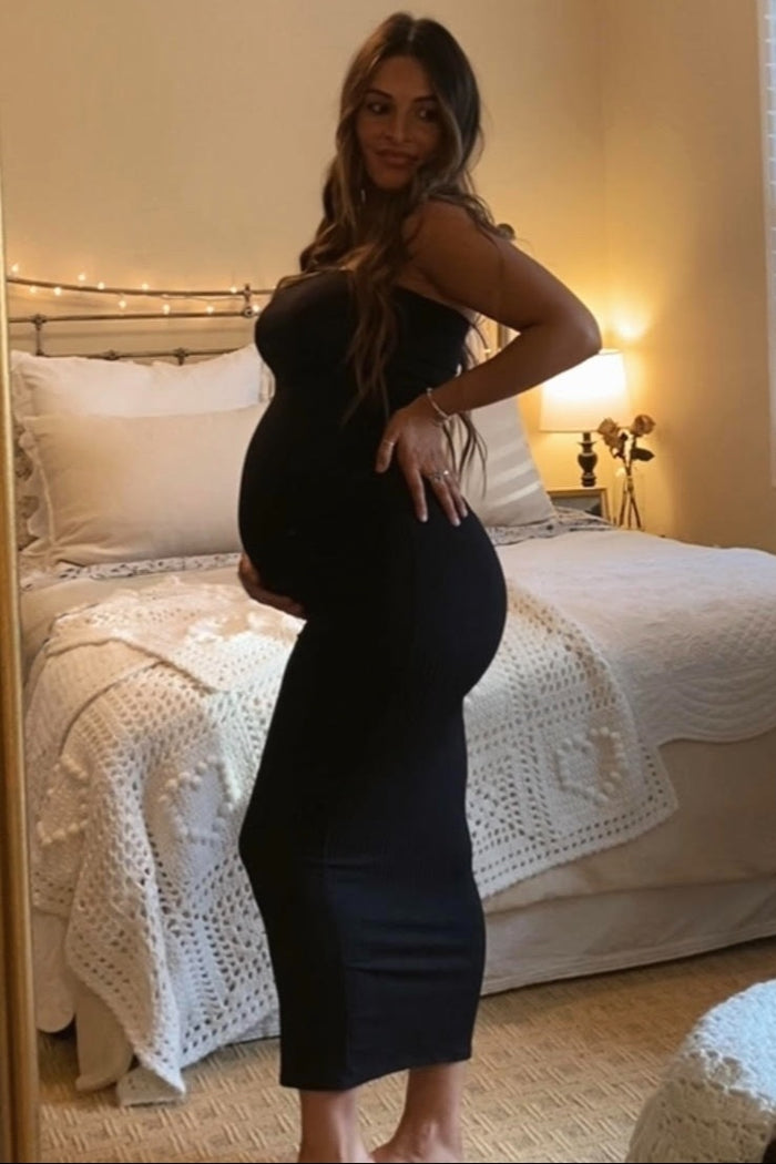 woman wearing a black strapless maternity dress, showcasing her baby bump, standing in a softly lit bedroom
