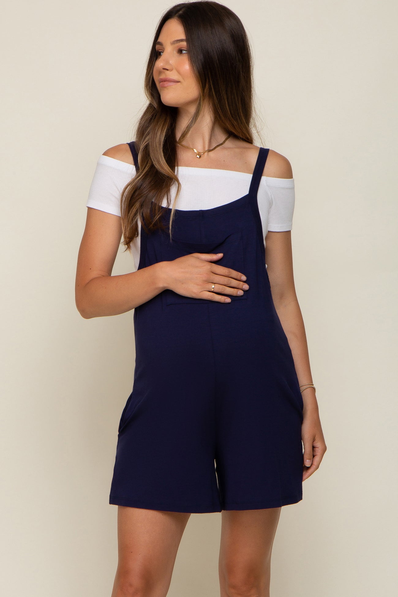 woman wearing a navy blue maternity romper with an off-shoulder white top and holding her baby bump