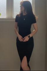 woman wearing a black fitted dress with a side slit, standing by a window with soft lighting