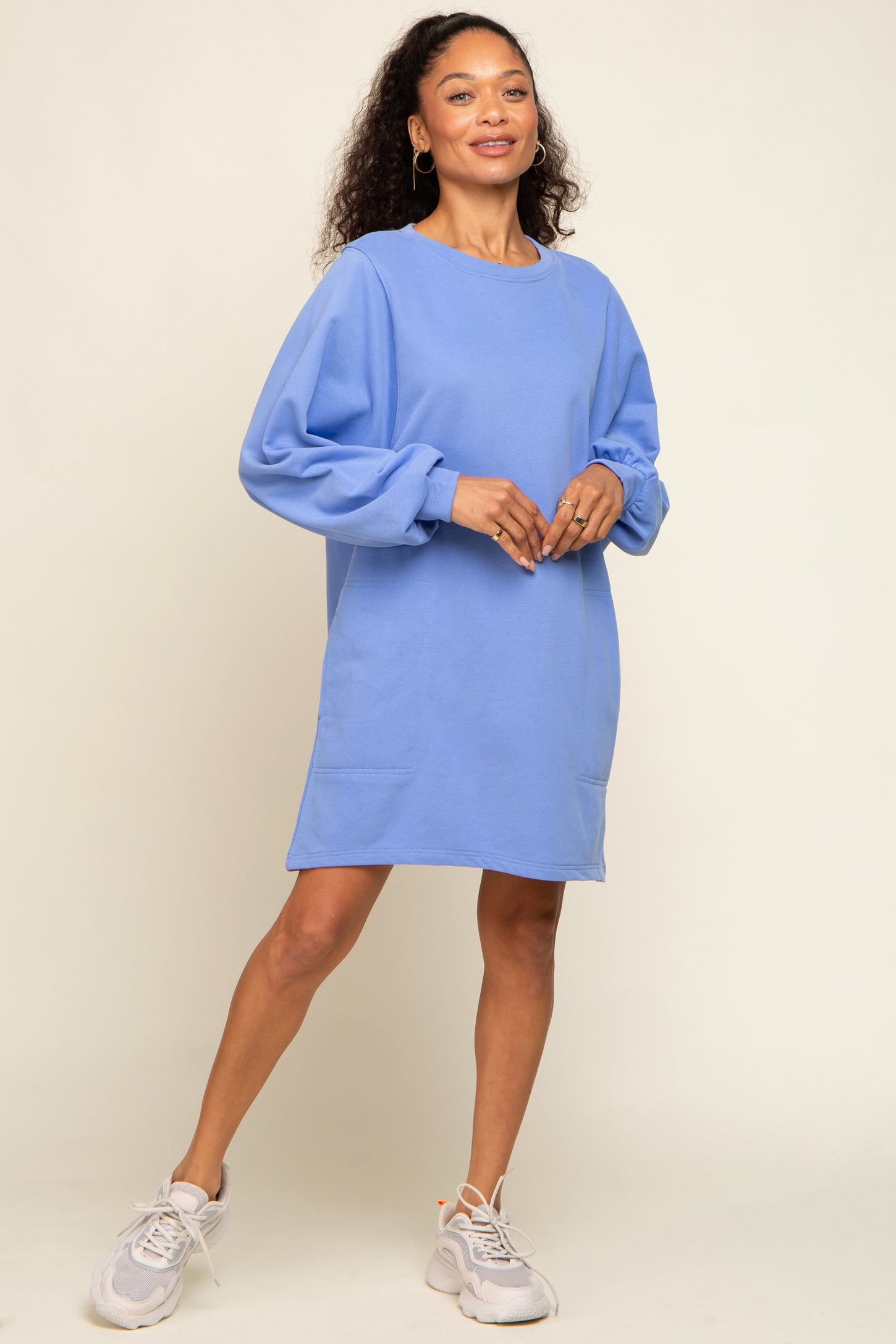 woman wearing a light blue oversized sweatshirt dress with balloon sleeves and pockets, standing on a neutral background