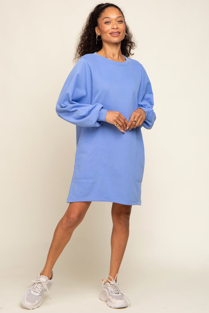 woman wearing a light blue oversized sweatshirt dress with balloon sleeves and pockets, standing on a neutral background