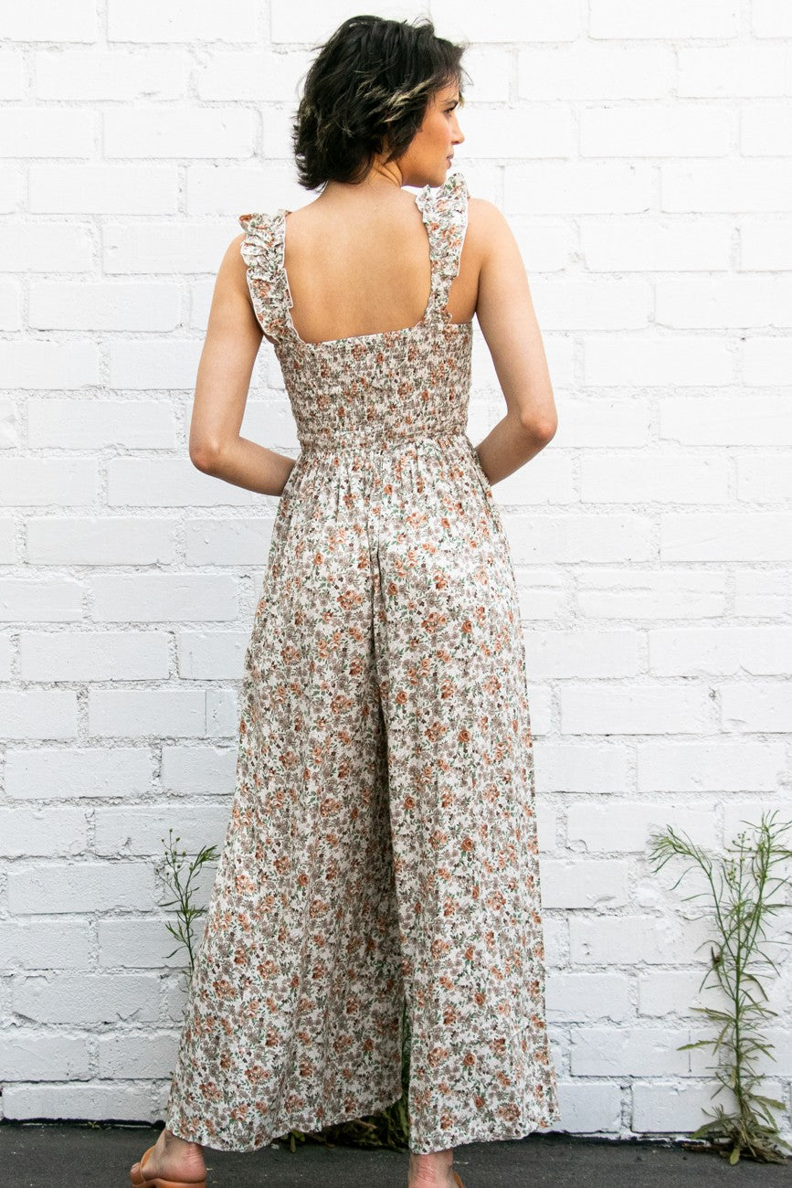 woman wearing a floral print jumpsuit with ruffled straps and a smocked back, standing against a white brick wall