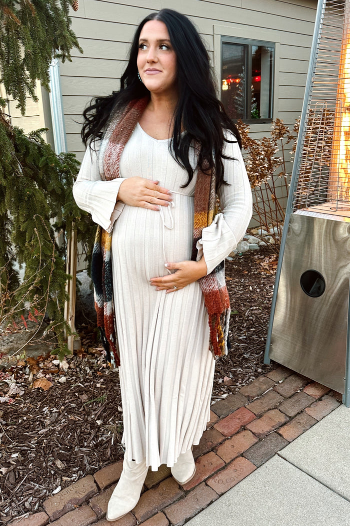 woman wearing a light beige maternity dress with long sleeves and a tie at the waist, holding her baby bump while standing outdoors