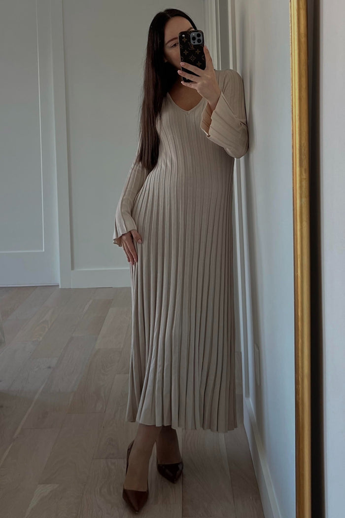 woman wearing a beige ribbed maxi dress with long sleeves and a deep v-neck, standing against a wall