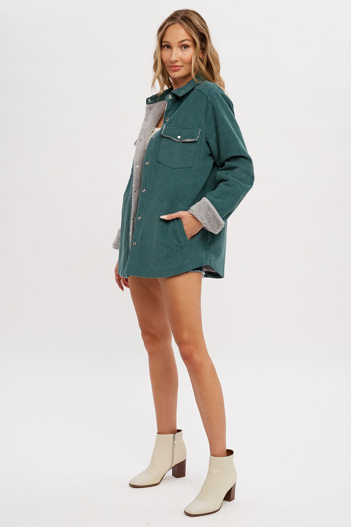 woman wearing a green corduroy oversized shirt jacket with gray fleece lining and pockets, paired with white ankle boots