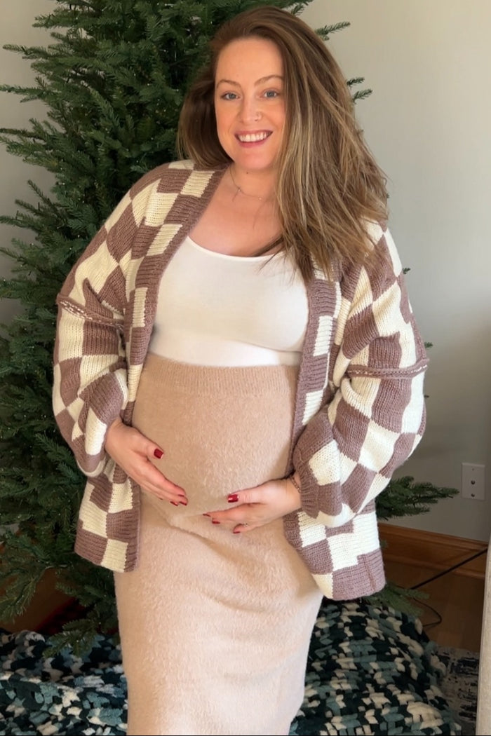 woman wearing a cream and brown checkered maternity cardigan and a beige maternity skirt, holding her baby bump