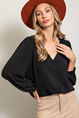 woman wearing a black v-neck blouse with puff sleeves and a tan hat, posing with one hand on her hip