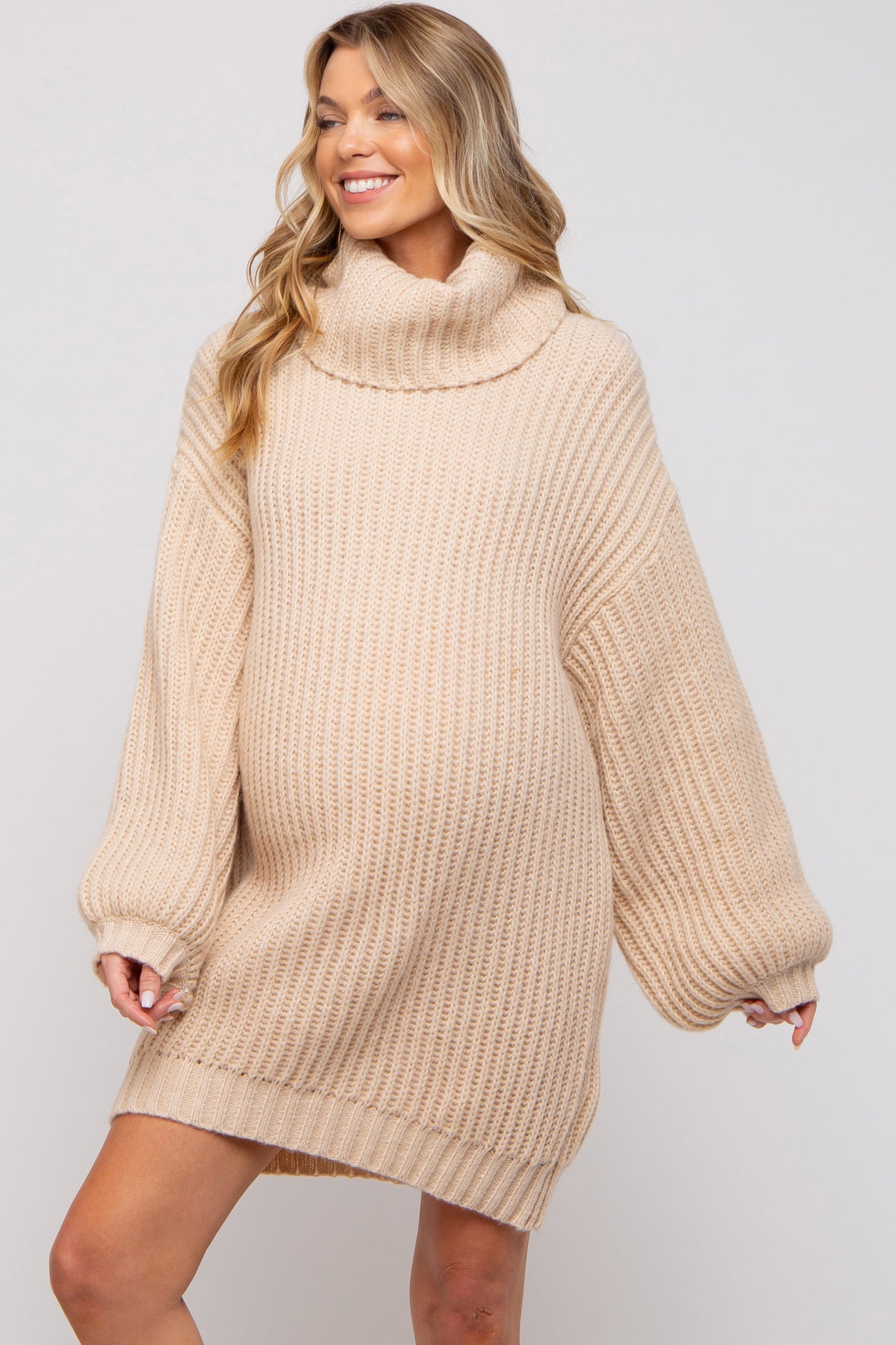 woman wearing a beige chunky knit maternity sweater dress with a turtleneck and oversized sleeves, showcasing her baby bump