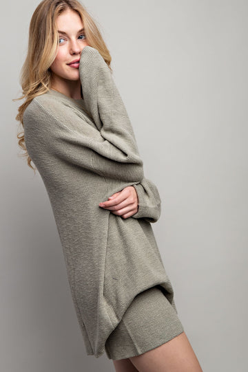 woman wearing a light olive green oversized sweater and matching shorts, smiling and posing sideways
