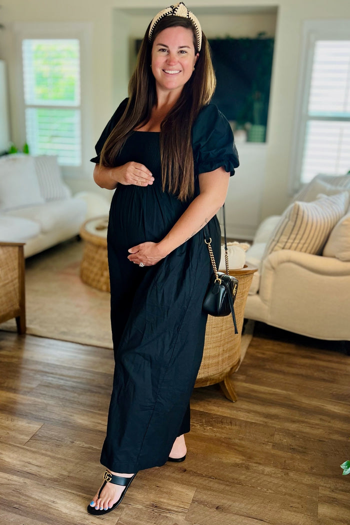 woman wearing a black maternity maxi dress and posing with her baby bump, accessorized with a headband and bag