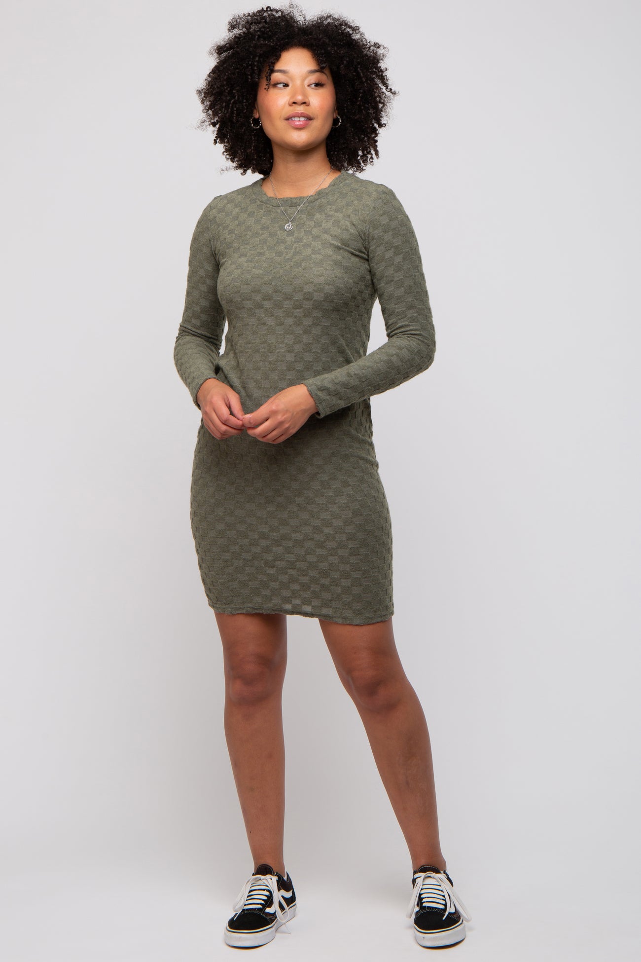 woman wearing an olive green long sleeve fitted dress with a textured checkered pattern and black sneakers