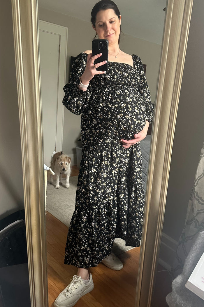 woman wearing a black floral maternity maxi dress and holding her baby bump with a dog in the background