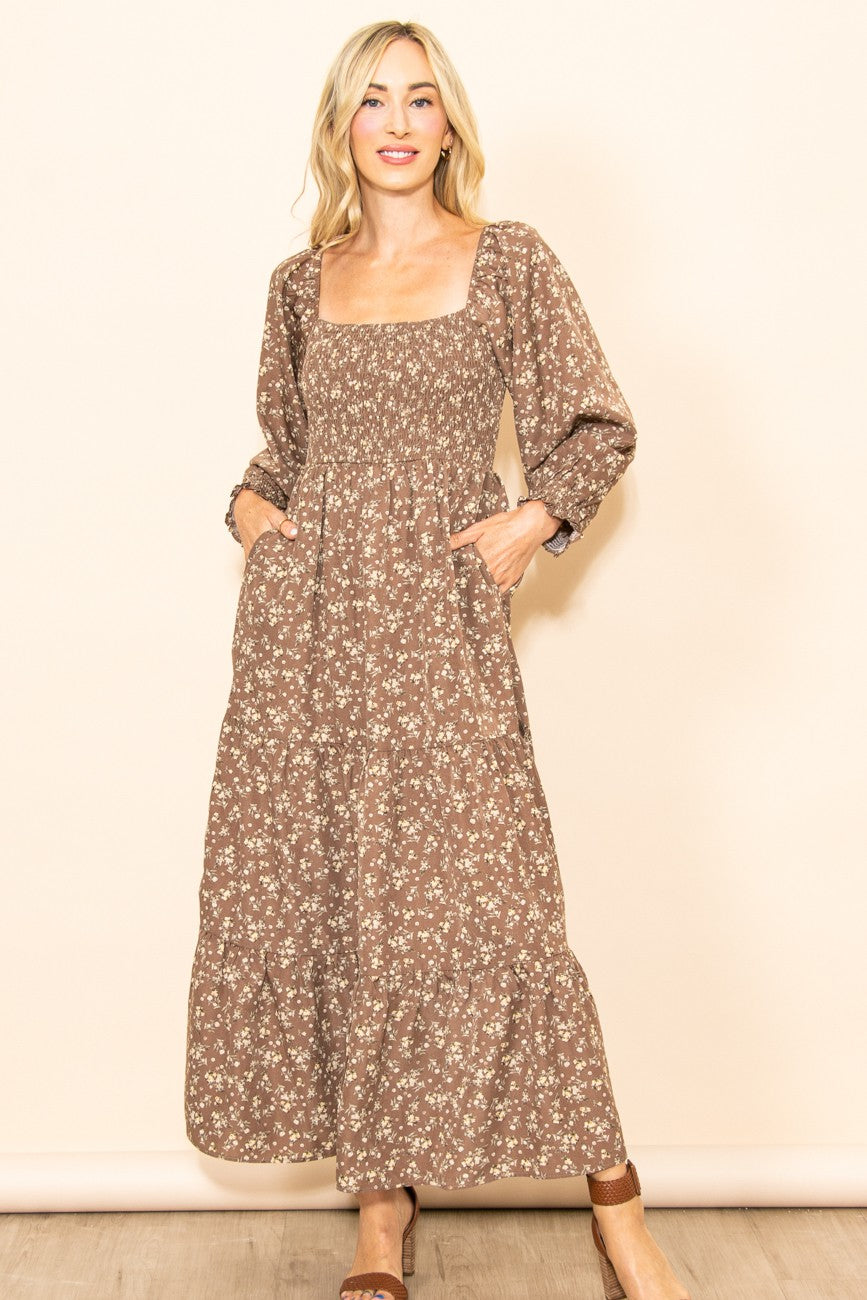 woman wearing a brown floral maxi dress with a square neckline and long sleeves, standing with hands in pockets