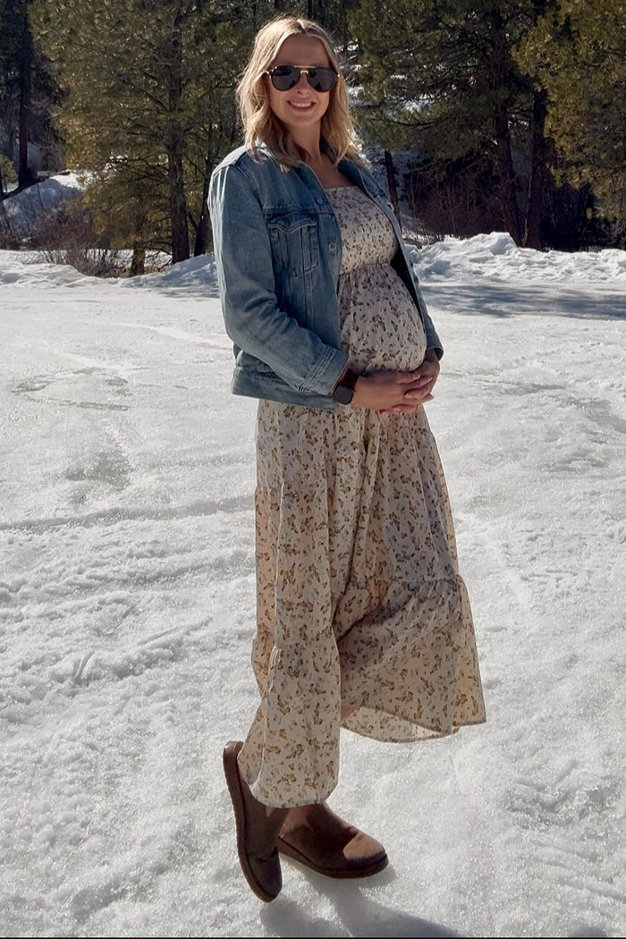 woman wearing a floral off-white maternity dress and a denim jacket, holding her baby bump in a snowy setting