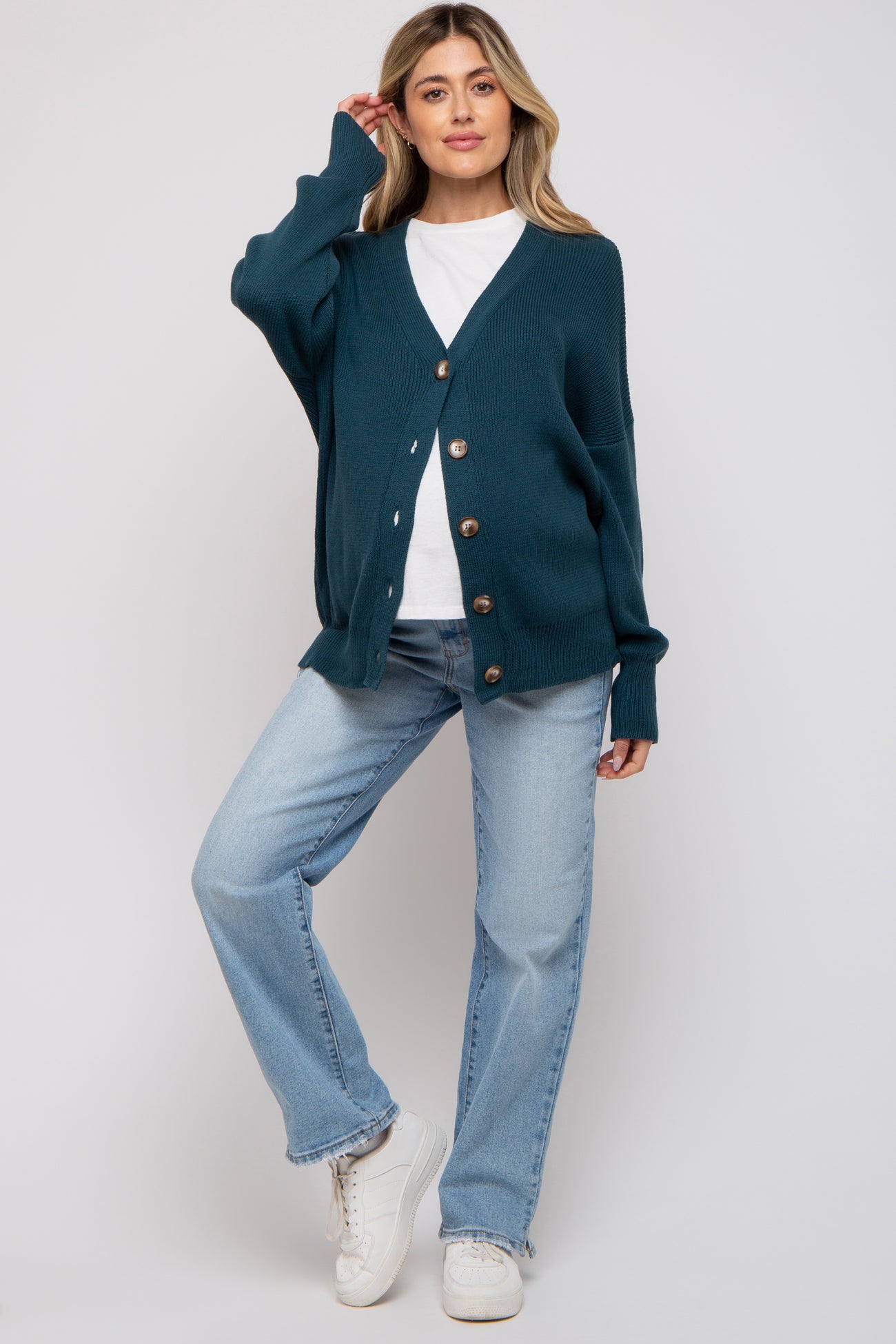 woman wearing a teal maternity cardigan over a white top and light blue jeans, standing in a casual pose