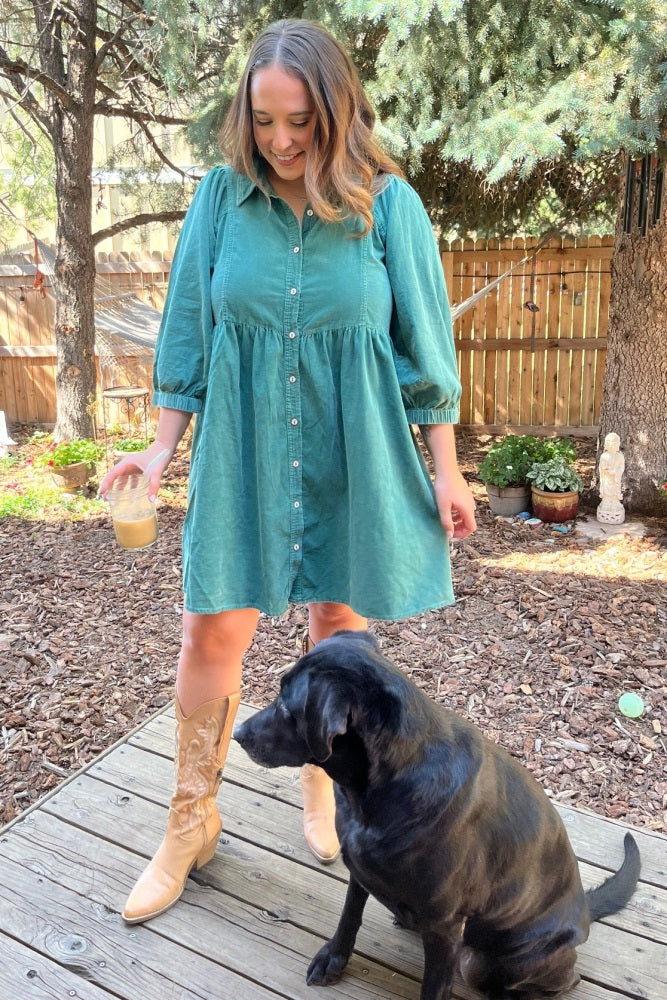 woman wearing a teal button-up maternity dress with puff sleeves and holding a drink, standing beside a black dog