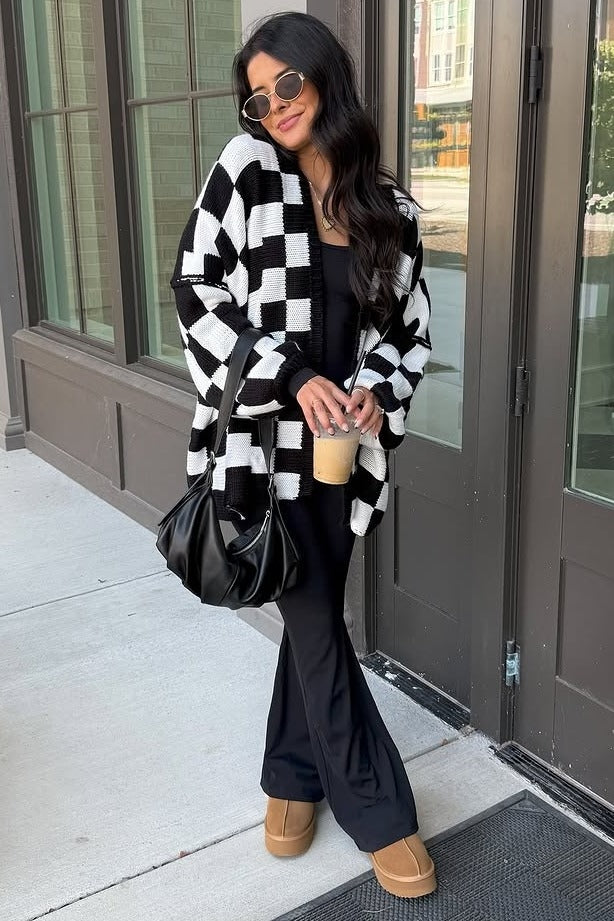 woman wearing a black and white checkered cardigan over a black outfit, holding a drink, standing outside