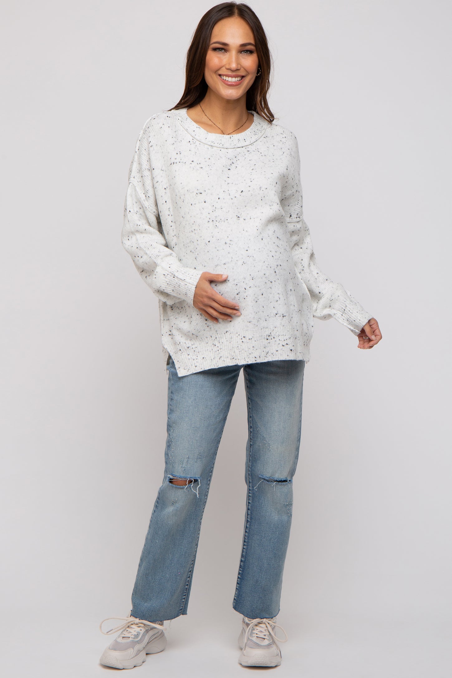 woman wearing a white speckled maternity sweater and distressed jeans, holding her baby bump