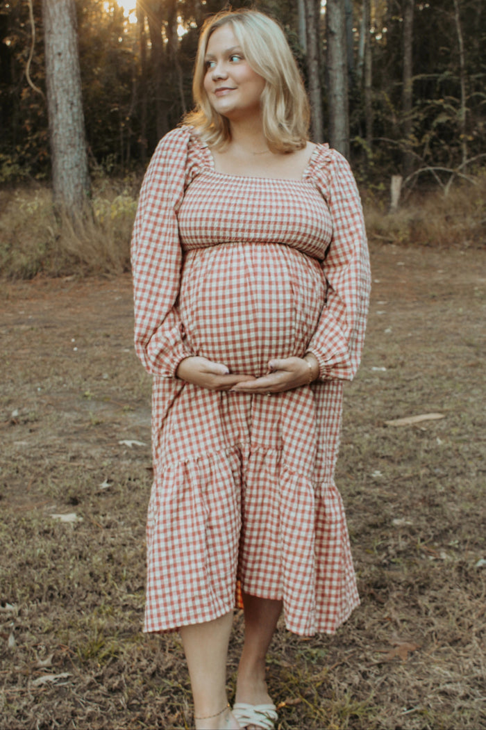 woman wearing a pink and white gingham maternity dress with long sleeves, holding her baby bump in a forest setting