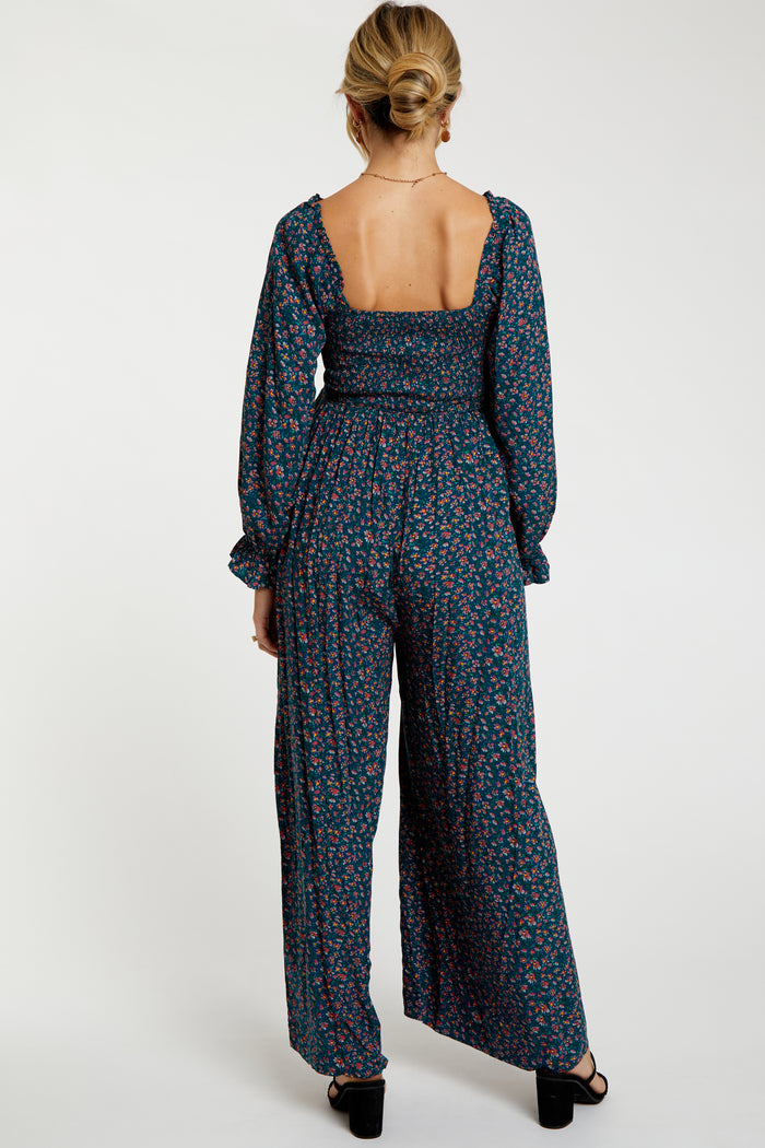 woman wearing a dark floral maternity jumpsuit with a smocked bodice and long sleeves, shown from the back