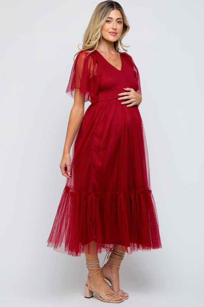 woman wearing a maroon maternity dress with sheer flutter sleeves and a tiered skirt, holding her baby bump