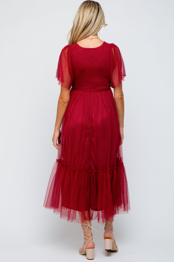 woman wearing a burgundy maternity dress with sheer sleeves and a tiered tulle skirt, showing a baby bump from the back