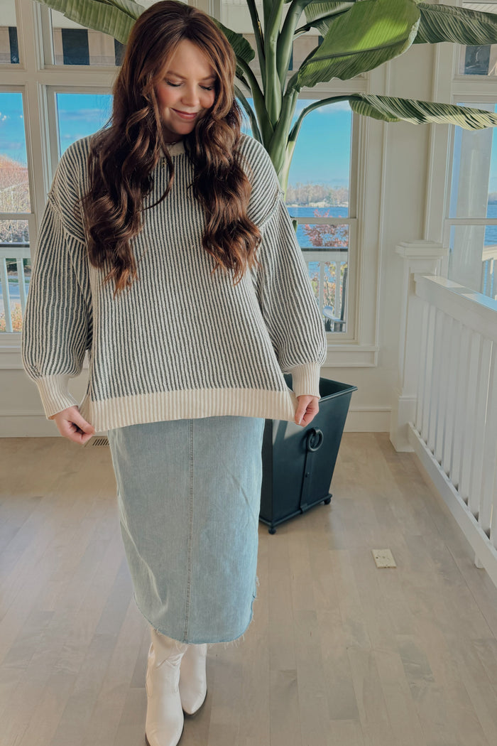 woman wearing a gray and white striped oversized sweater paired with a light denim midi skirt and white knee-high boots