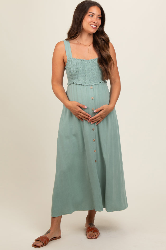 woman wearing a sage green maternity dress with smocked bodice and button details, cradling her baby bump