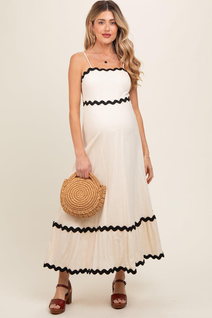 woman wearing a cream maternity maxi dress with black ruffled trim and holding a straw handbag