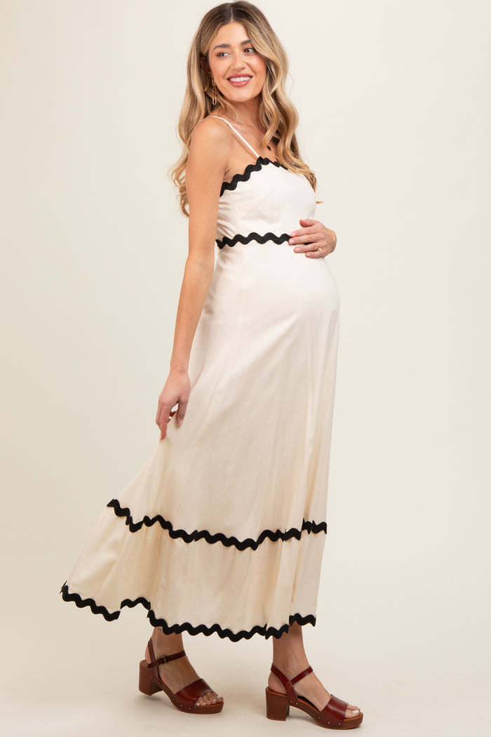 woman wearing a cream maternity maxi dress with black ric-rac trim and holding her baby bump