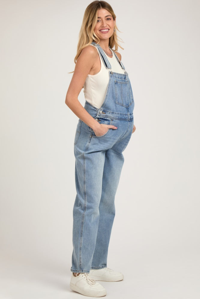 woman wearing light blue maternity overalls with pockets, paired with a fitted white tank top and white sneakers.