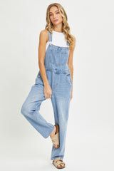 woman wearing light blue denim overalls over a white tank top, posing with one leg forward and wearing beige sandals