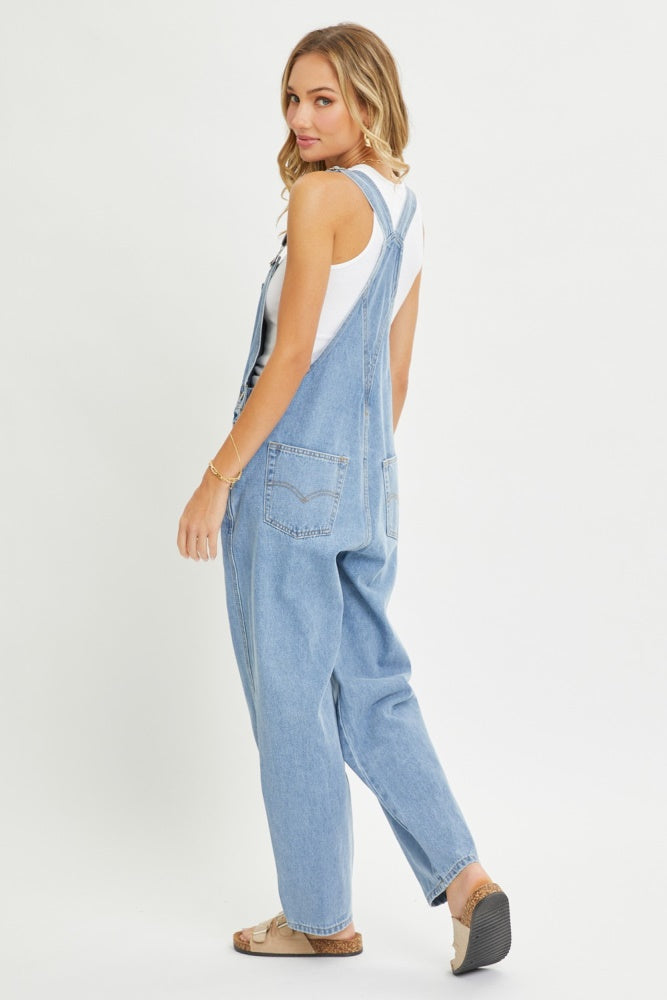 woman wearing a light blue denim overall with a white tank top, showing a relaxed fit and back view of the garment
