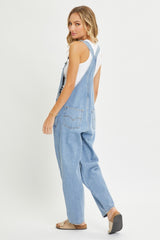 woman wearing a light blue denim overall with a white tank top, showing a relaxed fit and back view of the garment