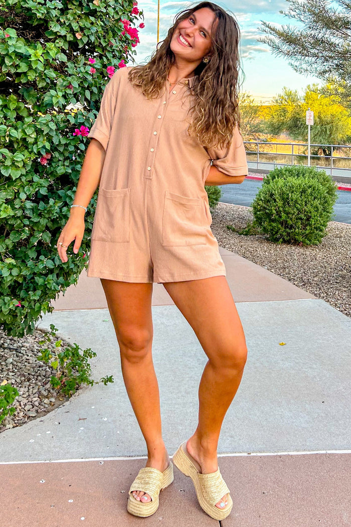 woman wearing a tan short sleeve romper with buttons and pockets, paired with wedge sandals and standing outside