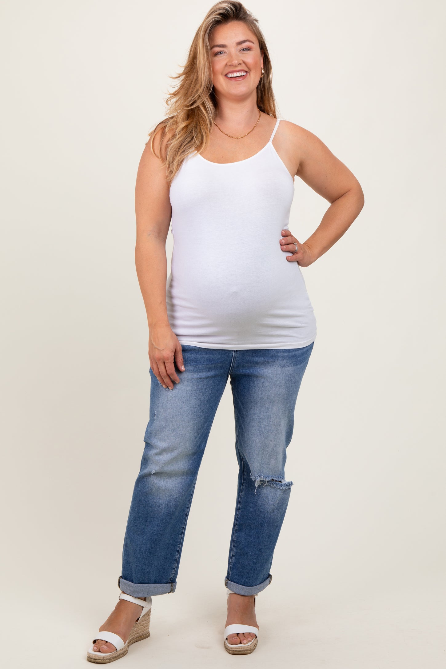 woman wearing a white maternity tank top and blue distressed jeans, showcasing her baby bump, standing with one hand on her hip
