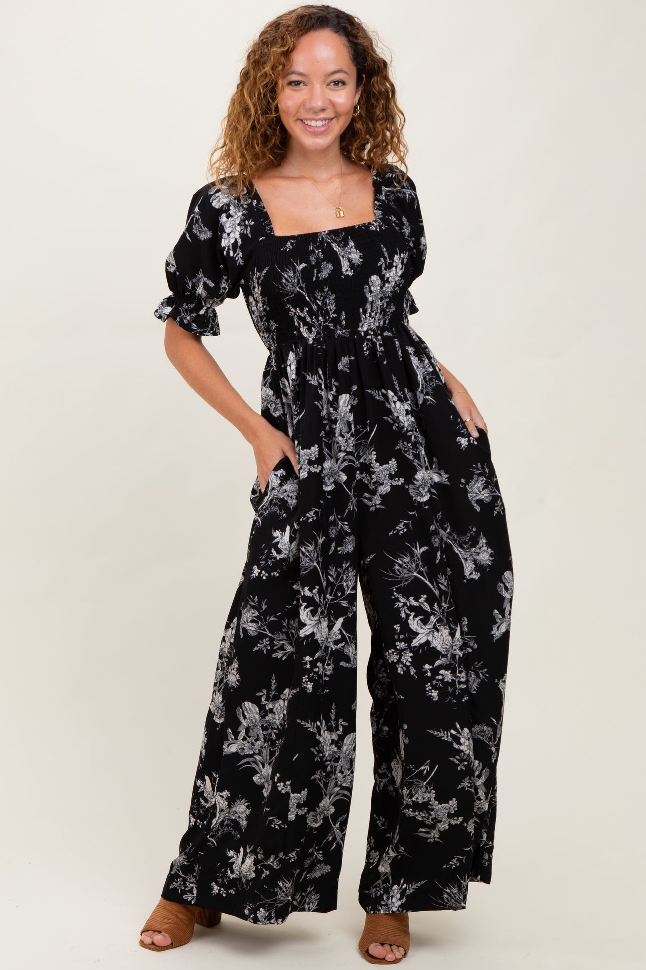 woman wearing a black floral print jumpsuit with puff sleeves and pockets, smiling at the camera