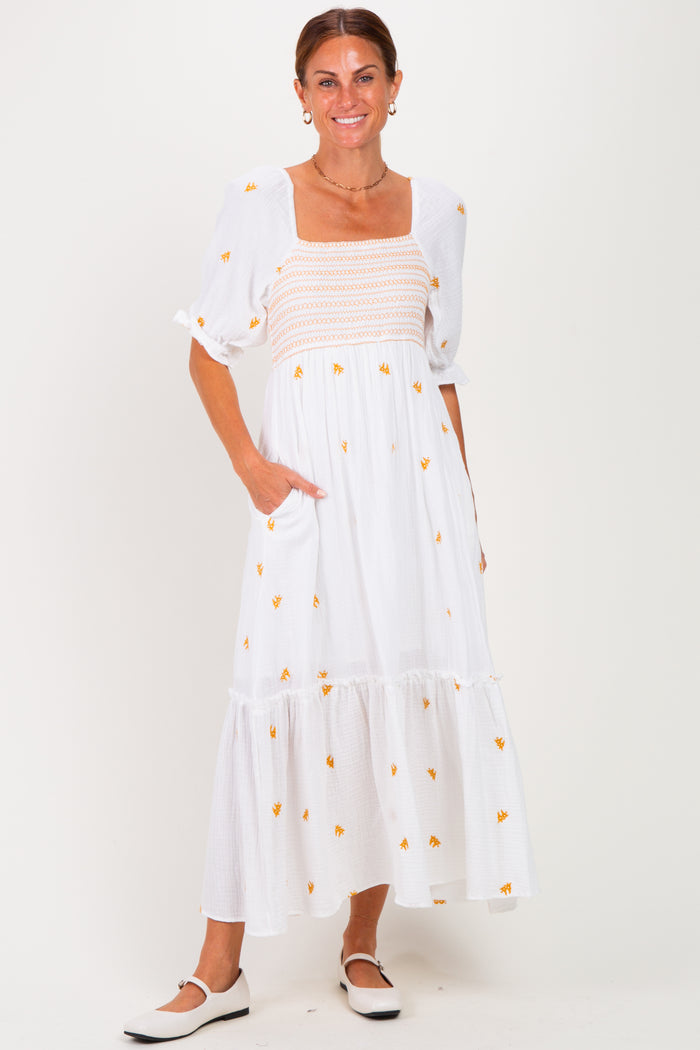 woman wearing a white maxi dress with yellow floral embroidery and puff sleeves, standing with hands in pockets