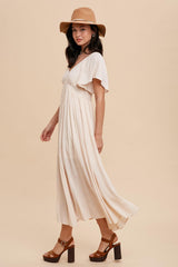 woman wearing a light beige maxi dress with flutter sleeves, paired with a tan hat and brown block-heeled sandals