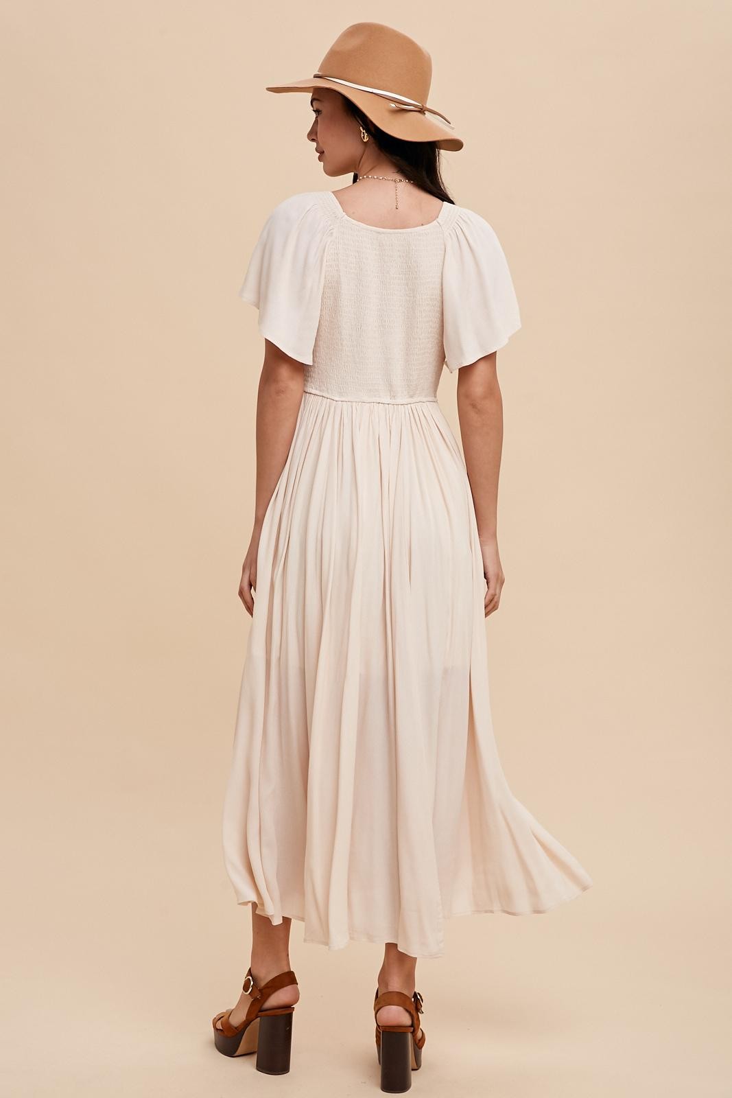 woman wearing a cream backless maxi dress with flutter sleeves and a smocked bodice, paired with a tan hat and heeled sandals