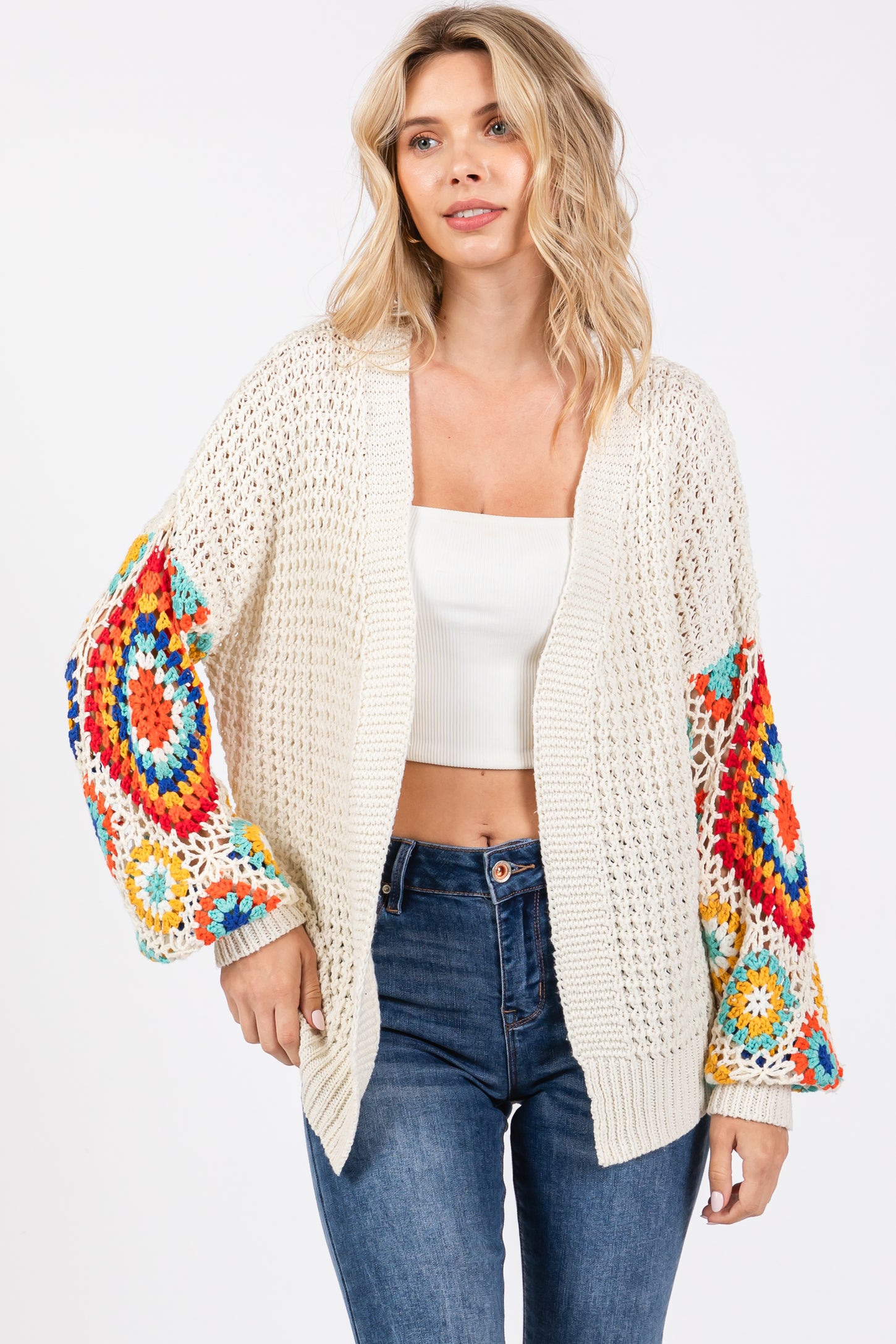 woman wearing a cream crochet cardigan with colorful patterned sleeves and a white crop top, paired with blue jeans