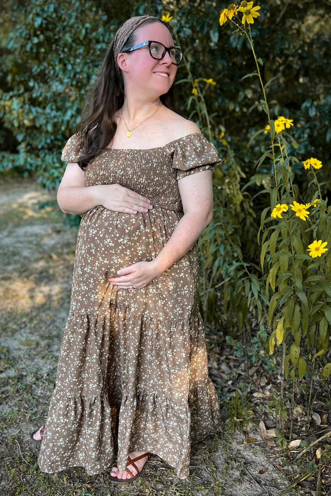 woman wearing a brown floral off-shoulder maternity dress and holding her baby bump in an outdoor setting